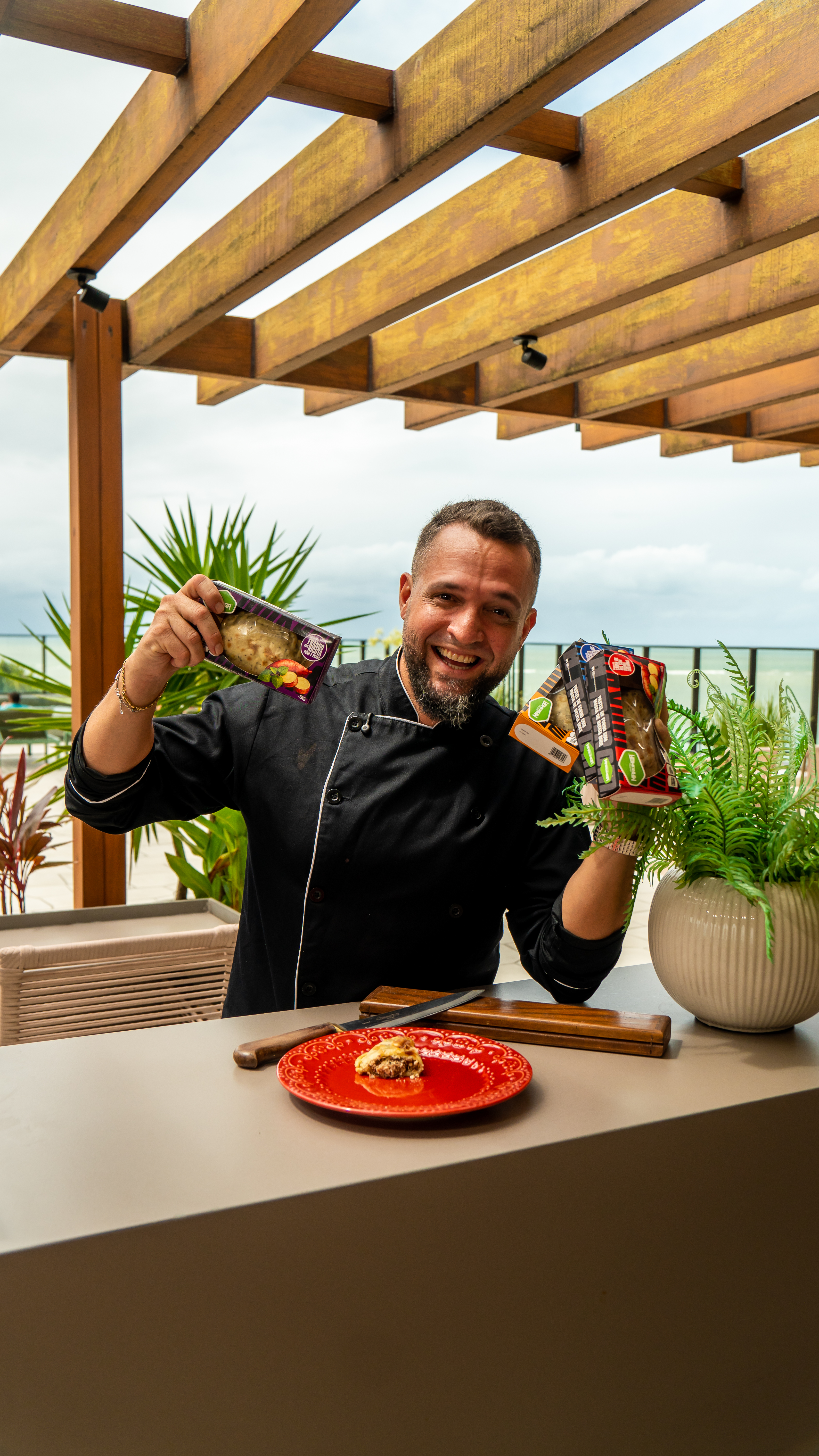 Homem de dólmã preta sorrindo e segurando duas caixas de Pastel Maromba, com um pastel cortado em um prato vermelho sobre a mesa